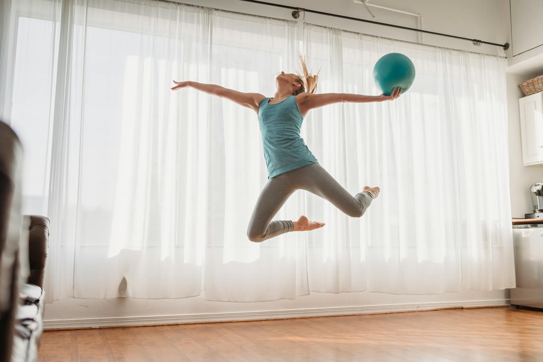 אימון בטן בבית — Young female in sportswear jumping up with bent legs and holding fit ball in hand on background of white cur